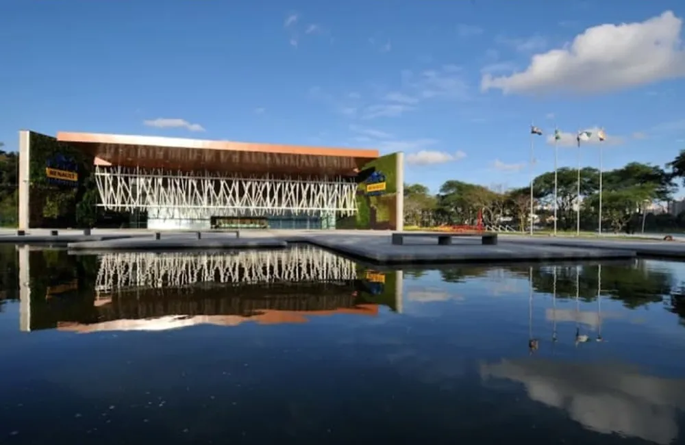 Fachada do Centro de Eventos Positivo refletida no espelho d'água, em dia de céu azul